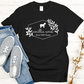 A short sleeve t-shirt by Amillia Acres ~ Goat Milk Soaps and Lotions displays a white goat graphic amidst leaves with "Goat Milk Soaps" text. Made from Gildan Softstyle 100% preshrunk cotton in black, it pairs effortlessly with blue jeans and gray sneakers, complemented by cotton stems nearby.