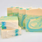 Amillia Acres' Awaken Goat Milk Soap, infused with eucalyptus and spearmint, stands upright against a light background. The handcrafted soap features marbled swirls of green, blue, and cream with specks of blue and green powder on two stacked bars.