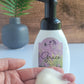 A hand holds Amillia Acres' Happiness Foaming Hand Soap with a white bottle and black pump. The purple label features lavender imagery and the word "Happiness." A blurred potted plant and wooden background appear in the distance.