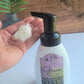 A hand holds "Relax" foaming hand soap from a white Amillia Acres bottle with lavender design against a wooden background and green plant, highlighting the goat milk's moisturizing benefits.